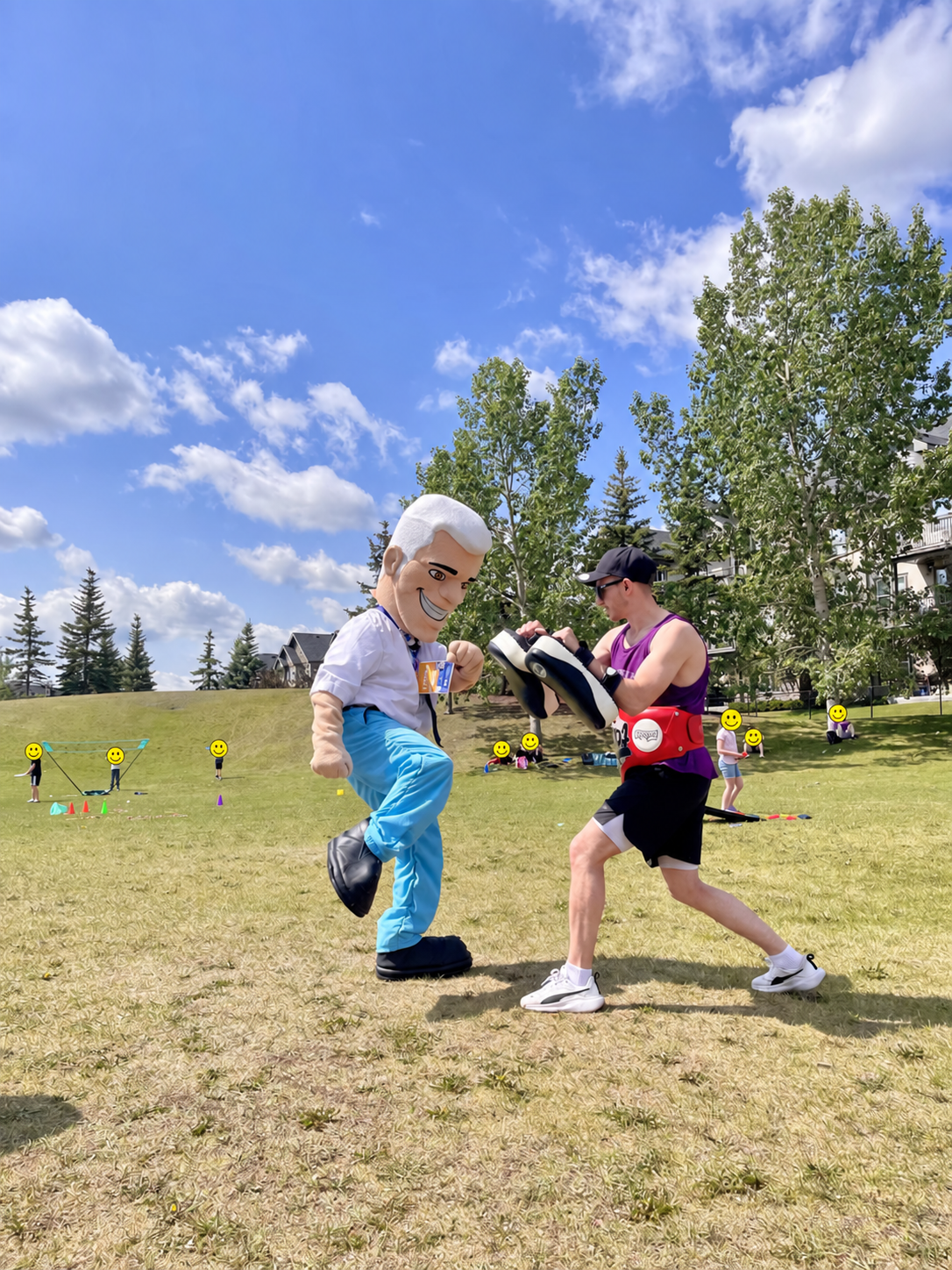 Logan practising Muay Thai padwork with a mascot at an outdoor Calgary community event, with children's faces covered by smiley graphics