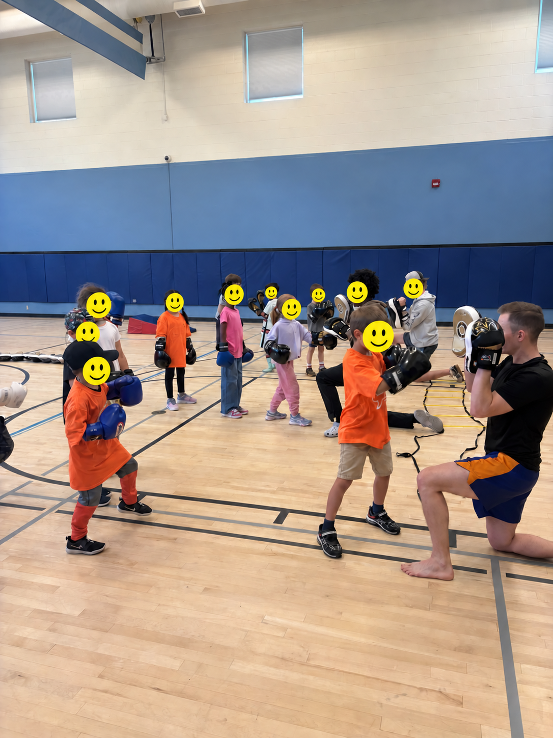 Children and teen pad holders practising Muay Thai during a community gym program, with every minor's face covered by a smiley graphic